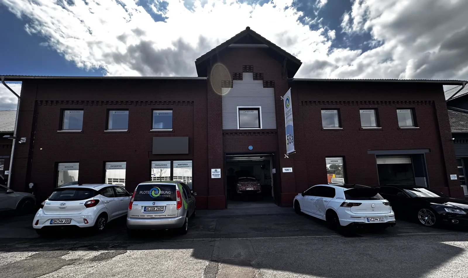 Backstein-Firmengebäude mit Werkstatt-Tor und parkenden Autos unter Wolkenhimmel